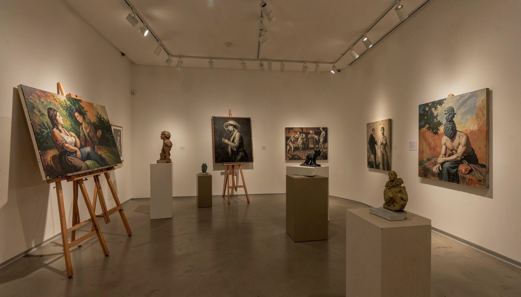 An intimate gallery space with track lighting, featuring a mix of paintings on easels and sculptural works on pedestals in muted earth tones.